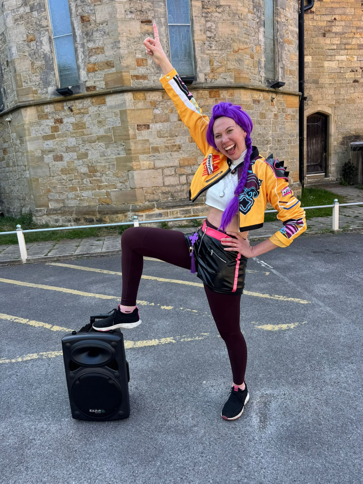 Anna in a colourful yellow jacket and purple wig, striking a party pose beside her speaker